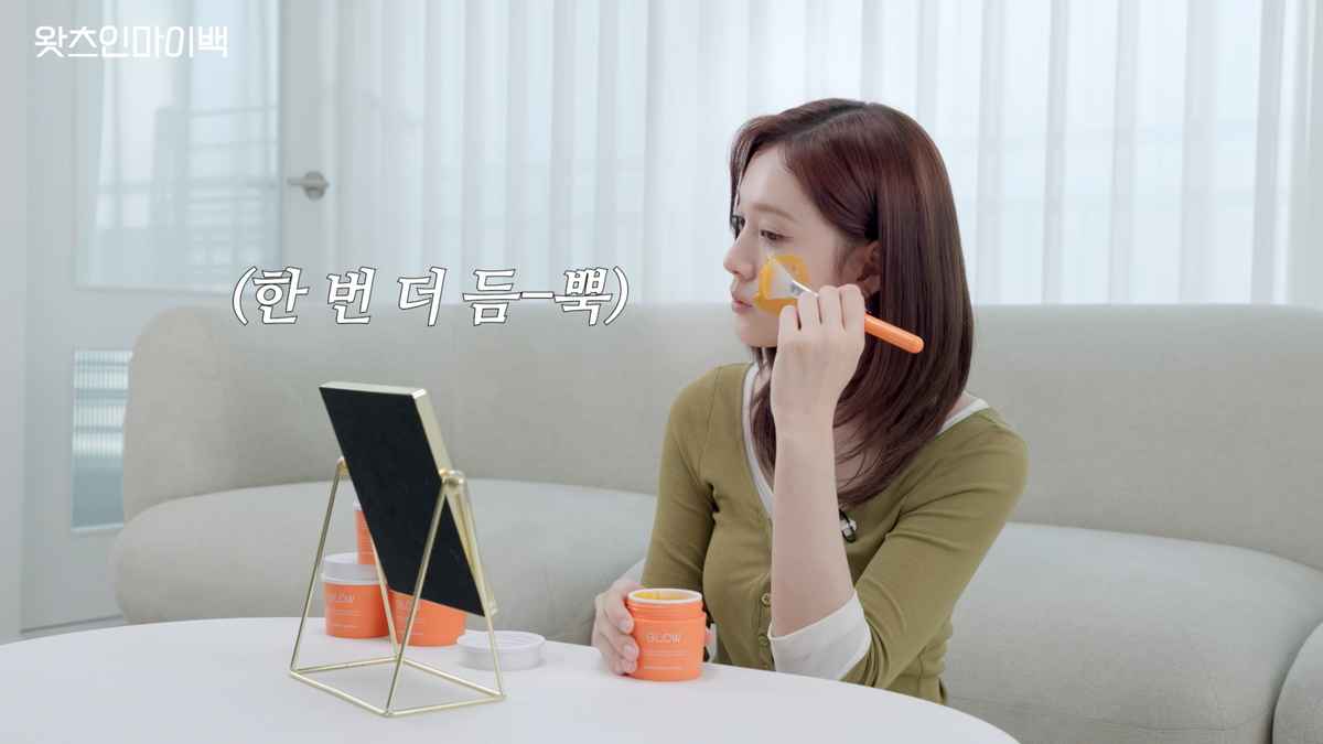 STAYC J applying FAU Glow Pumpkin 7.0 Acid Peel skincare mask to her cheek with a brush in front of a mirror.